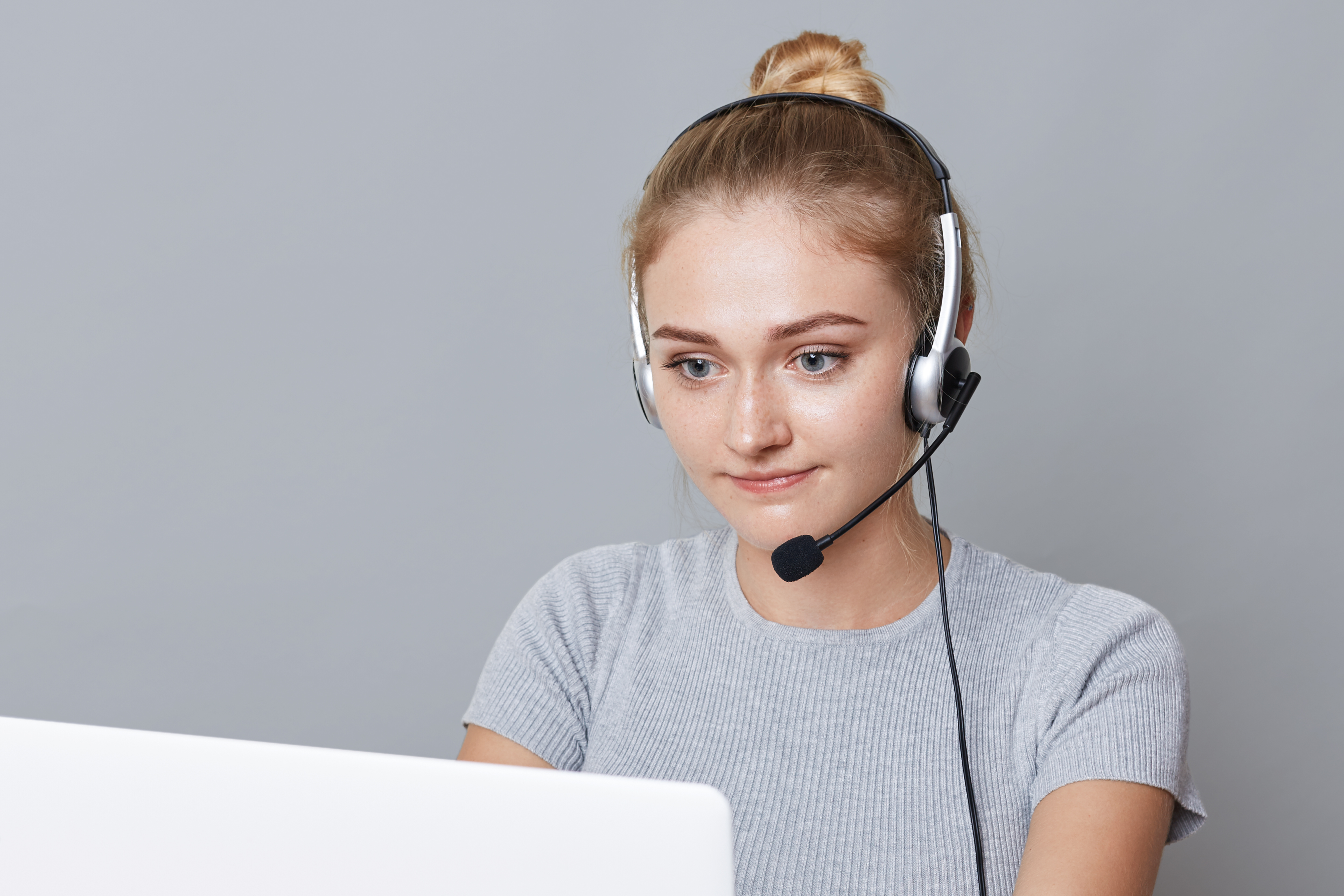 Portrait of confident female with hair knot looks makes video call, sits in front of opened laptop, communicates with friends, isolated over grey background. Young woman has online conference